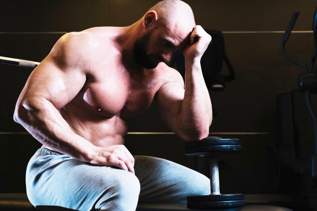 Bodybuilder-reflecting-while-sitting-in-gym-with-dumbbell